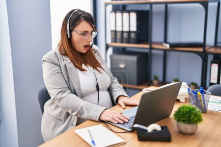 Foto de Mujer embarazada que trabaja en la oficina con los auriculares del operador asustado y sorprendido con la boca abierta para sorpresa, cara de incredulidad - Imagen libre de derechos