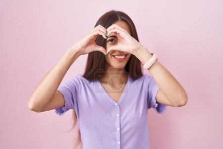 Foto de Mujer hispana joven con el pelo largo de pie sobre fondo rosa haciendo forma de corazón con la mano y los dedos sonriendo mirando a través de signo - Imagen libre de derechos