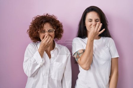 Foto de Madre e hija hispanas usando una camiseta blanca casual sobre fondo rosa oliendo algo apestoso y asqueroso, olor intolerable, conteniendo la respiración con los dedos en la nariz. mal olor - Imagen libre de derechos