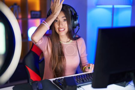 Photo for Young hispanic woman playing video games surprised with hand on head for mistake, remember error. forgot, bad memory concept. - Royalty Free Image