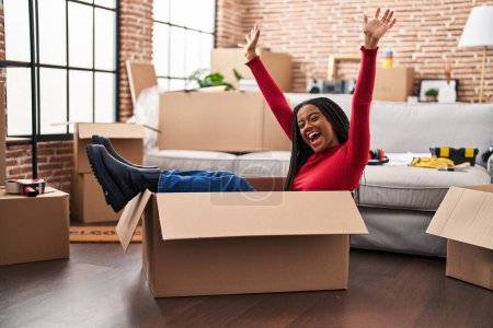 Foto de Joven afroamericano con trenzas mudándose a un nuevo hogar dentro de una caja de cartón celebrando la victoria con sonrisa feliz y expresión ganadora con las manos levantadas - Imagen libre de derechos