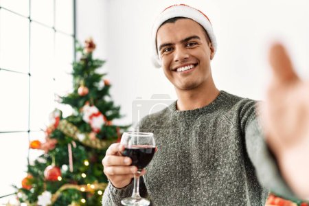 Téléchargez les photos : Jeune homme hispanique faire selfie par la caméra boire du vin debout près de l'arbre de Noël à la maison - en image libre de droit