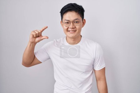 Téléchargez les photos : Young asian man standing over white background smiling and confident gesturing with hand doing small size sign with fingers looking and the camera. measure concept. - en image libre de droit