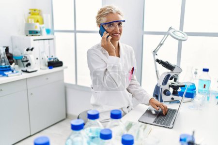 Photo for Middle age blonde woman wearing scientist uniform talking on the smartphone working at laboratory - Royalty Free Image