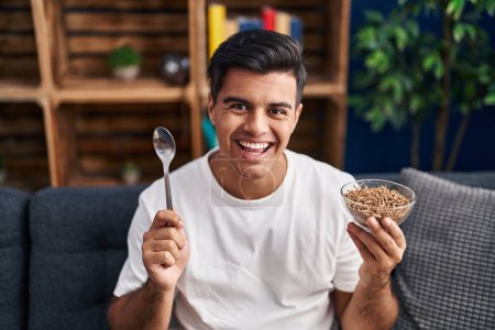 Photo for Hispanic man eating healthy whole grain cereals with spoon smiling and laughing hard out loud because funny crazy joke. - Royalty Free Image