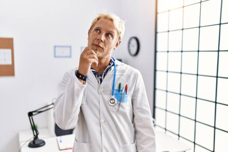 Photo for Young blond man wearing doctor uniform and stethoscope at clinic with hand on chin thinking about question, pensive expression. smiling with thoughtful face. doubt concept. - Royalty Free Image