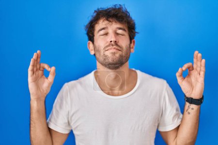Photo for Hispanic young man standing over blue background relaxed and smiling with eyes closed doing meditation gesture with fingers. yoga concept. - Royalty Free Image