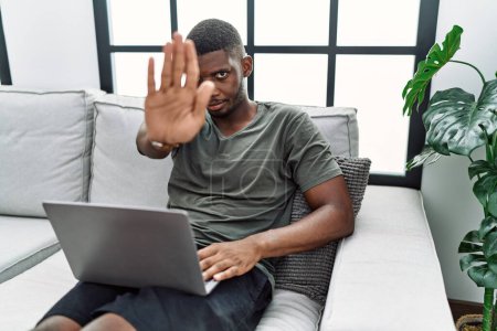 Photo for Young african american man using laptop at home sitting on the sofa doing stop sing with palm of the hand. warning expression with negative and serious gesture on the face. - Royalty Free Image