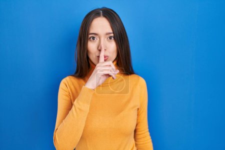Photo for Young brunette woman standing over blue background asking to be quiet with finger on lips. silence and secret concept. - Royalty Free Image