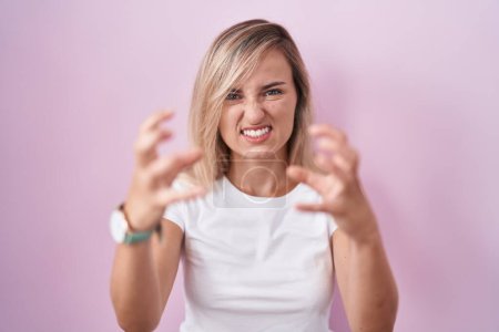 Photo for Young blonde woman standing over pink background shouting frustrated with rage, hands trying to strangle, yelling mad - Royalty Free Image