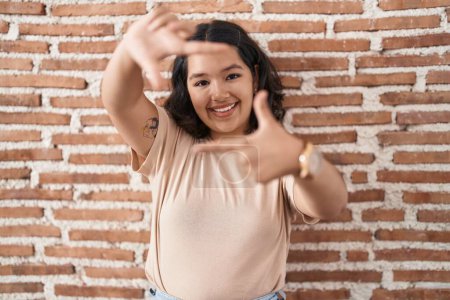 Foto de Mujer hispana joven de pie sobre la pared de ladrillos sonriendo haciendo marco con las manos y los dedos con la cara feliz. concepto de creatividad y fotografía. - Imagen libre de derechos