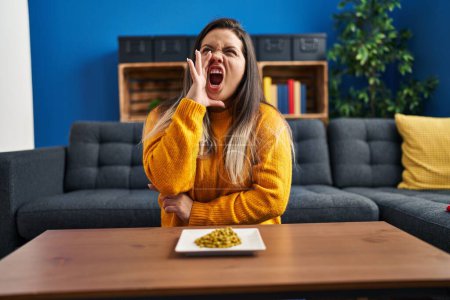 Photo for Young hispanic woman eating green peas clueless and confused with open arms, no idea and doubtful face. - Royalty Free Image