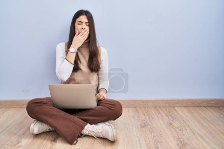 Téléchargez les photos : Jeune femme brune travaillant à l'aide d'un ordinateur portable assis sur le sol ennuyé bâillement fatigué couvrant la bouche avec la main. agitation et somnolence. - en image libre de droit