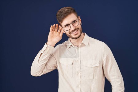 Foto de Hombre hispano con barba de pie sobre fondo azul sonriendo con la mano sobre el oído escuchando un rumor o chismes. concepto de sordera. - Imagen libre de derechos