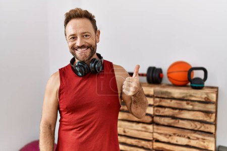Téléchargez les photos : Homme d'âge moyen portant des vêtements de sport et des écouteurs à la salle de gym souriant heureux et positif, pouce levé faire excellent et signe d'approbation - en image libre de droit