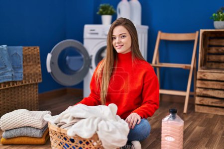 Foto de Young blonde woman folding clothes sitting on floor at laundry room - Imagen libre de derechos