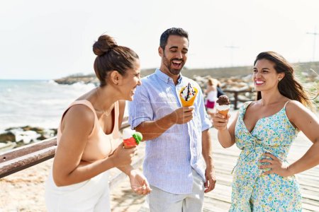Foto de Tres jóvenes amigos hispanos sonriendo felices comiendo helado en la playa. - Imagen libre de derechos