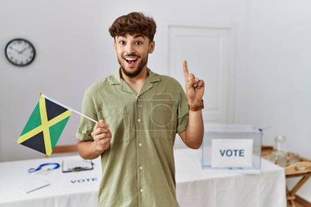 Foto de Young arab man at political campaign election holding jamaica flag surprised with an idea or question pointing finger with happy face, number one - Imagen libre de derechos