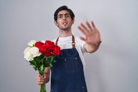 Foto de Joven hombre hispano sosteniendo un ramo de rosas blancas y rojas haciendo un gesto de stop con palmas de las manos, expresión de enojo y frustración - Imagen libre de derechos