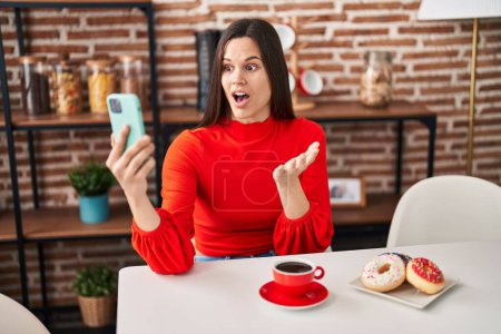 Foto de Young hispanic woman eating doughnuts and drinking a cup of coffee doing video call scared and amazed with open mouth for surprise, disbelief face - Imagen libre de derechos