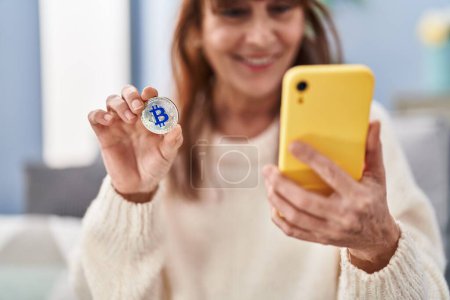 Photo for Middle age woman using smartphone holding bitcoin at home - Royalty Free Image