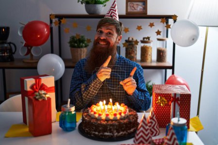 Foto de Hombre caucásico con barba larga celebrando cumpleaños sosteniendo gran pastel de chocolate sonriendo y mirando a la cámara apuntando con dos manos y dedos hacia un lado. - Imagen libre de derechos