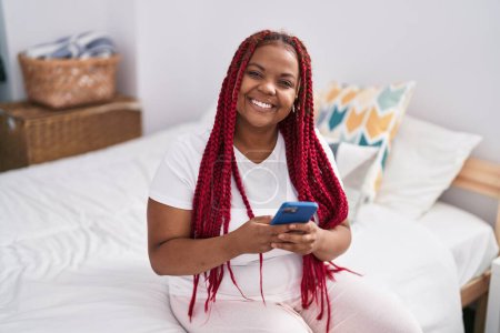 Téléchargez les photos : Femme afro-américaine utilisant smartphone assis sur le lit dans la chambre - en image libre de droit