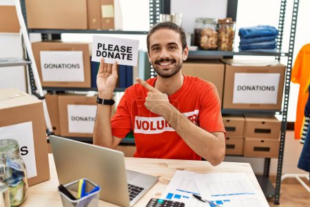 Photo for Young hispanic man wearing volunteer t shirt holding please donate banner smiling happy pointing with hand and finger - Royalty Free Image