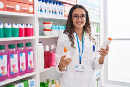 Foto de Joven hermosa mujer hispana farmacéutica sosteniendo botellas de crema en la farmacia - Imagen libre de derechos