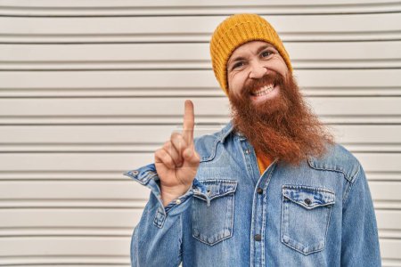 Téléchargez les photos : Homme caucasien avec longue barbe waring chapeau de laine souriant avec une idée ou une question pointant du doigt avec un visage heureux, numéro un - en image libre de droit