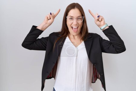 Photo for Beautiful brunette woman wearing business jacket and glasses smiling amazed and surprised and pointing up with fingers and raised arms. - Royalty Free Image