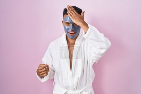 Téléchargez les photos : Young hispanic man wearing beauty face mask and bath robe surprised with hand on head for mistake, remember error. forgot, bad memory concept. - en image libre de droit