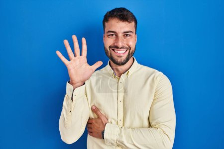 Photo for Handsome hispanic man standing over blue background showing and pointing up with fingers number five while smiling confident and happy. - Royalty Free Image