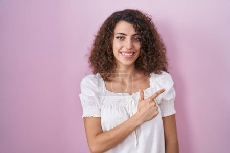 Foto de Mujer hispana con el pelo rizado de pie sobre fondo rosa alegre con una sonrisa de la cara señalando con la mano y el dedo hacia arriba a un lado con expresión feliz y natural en la cara - Imagen libre de derechos