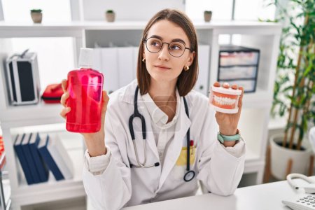 Téléchargez les photos : Young caucasian dentist woman holding denture and mouthwash smiling looking to the side and staring away thinking. - en image libre de droit