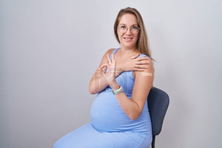 Foto de Mujer embarazada joven que usa una tirita para la inyección de vacunas haciendo buen signo con los dedos, sonriendo gesto amistoso excelente símbolo - Imagen libre de derechos