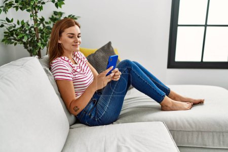 Téléchargez les photos : Jeune femme utilisant smartphone assis sur le canapé à la maison - en image libre de droit