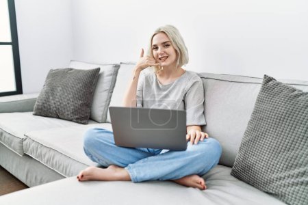 Photo for Young caucasian woman using laptop at home sitting on the sofa smiling doing phone gesture with hand and fingers like talking on the telephone. communicating concepts. - Royalty Free Image