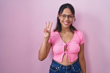 Téléchargez les photos : Hispanic young woman standing over pink background wearing glasses showing and pointing up with fingers number three while smiling confident and happy. - en image libre de droit