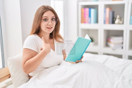 Foto de Joven mujer caucásica sentada en la cama en casa leyendo un libro señalando con el dedo a uno mismo sonriendo feliz y orgulloso - Imagen libre de derechos