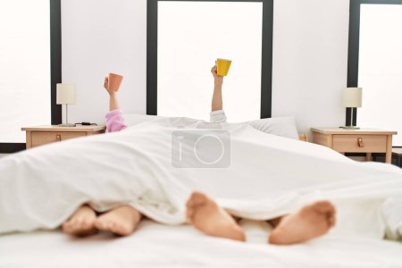 Téléchargez les photos : Jeune couple hispanique couché sur le lit avec une tasse soulevée dans la chambre. - en image libre de droit