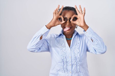 Foto de Mujer africana con rastas de pie sobre fondo blanco usando gafas haciendo buen gesto como prismáticos sacando la lengua, ojos mirando a través de los dedos. expresión loca. - Imagen libre de derechos