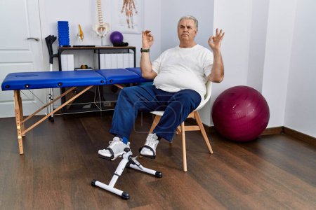 Photo for Senior caucasian man at physiotherapy clinic using pedal exerciser relax and smiling with eyes closed doing meditation gesture with fingers. yoga concept. - Royalty Free Image