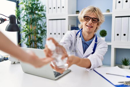 Photo for Young blond man doctor smiling confident using sanitizer gel hands at clinic - Royalty Free Image