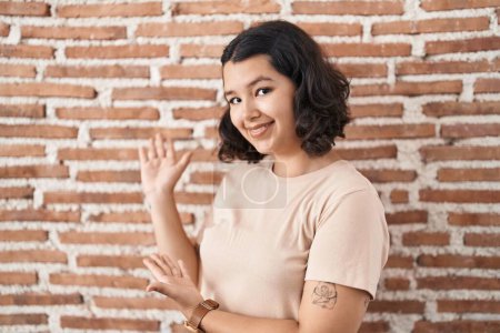 Foto de Mujer hispana joven de pie sobre la pared de ladrillos invitando a entrar sonriendo natural con la mano abierta - Imagen libre de derechos