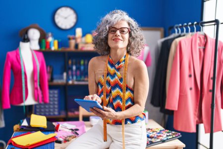 Téléchargez les photos : Middle age grey-haired woman tailor smiling confident using touchpad at sewing studio - en image libre de droit
