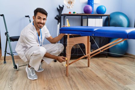 Photo for Young hispanic man wearing physiotherapist uniform manipulating massage table at rehab clinic - Royalty Free Image