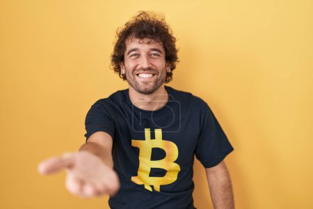 Téléchargez les photos : Jeune homme hispanique portant Bitcoin t-shirt souriant joyeuse offrant main de paume donnant assistance et acceptation. - en image libre de droit