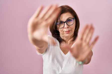 Foto de Mujer hispana de mediana edad de pie sobre fondo rosa haciendo marco usando las manos palmas y los dedos, perspectiva de la cámara - Imagen libre de derechos