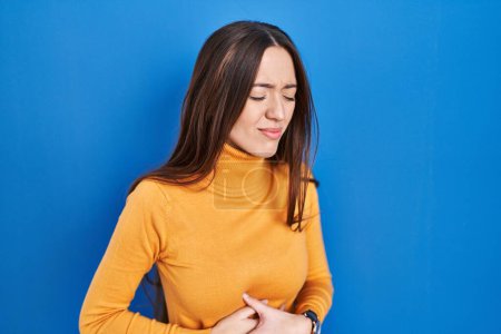 Photo for Young brunette woman standing over blue background with hand on stomach because indigestion, painful illness feeling unwell. ache concept. - Royalty Free Image